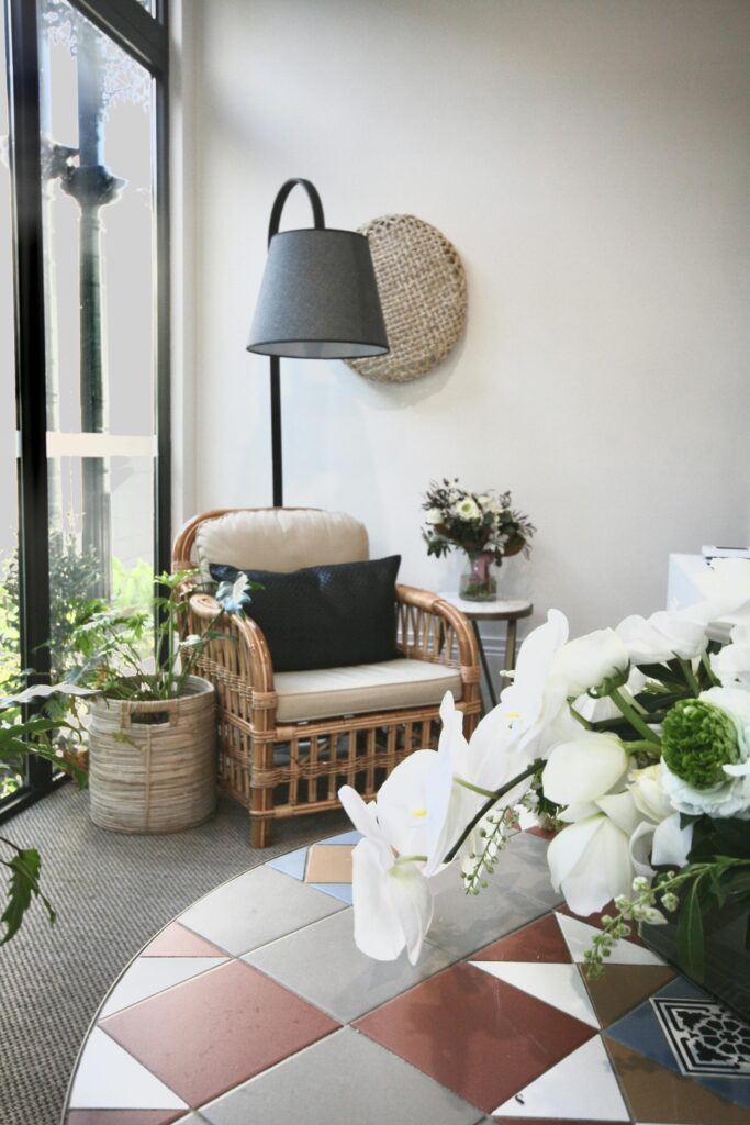 Poltrona de vime com almofada, iluminária cinza e vaso com flores.