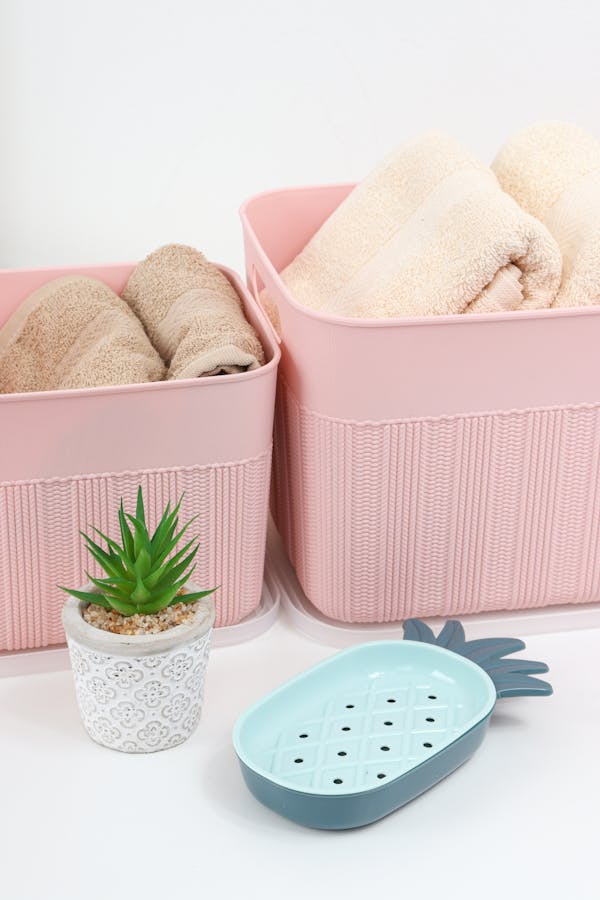 Caixas grande e média de plástico, cor de rosa, com toalhas para a organização do banheiro.