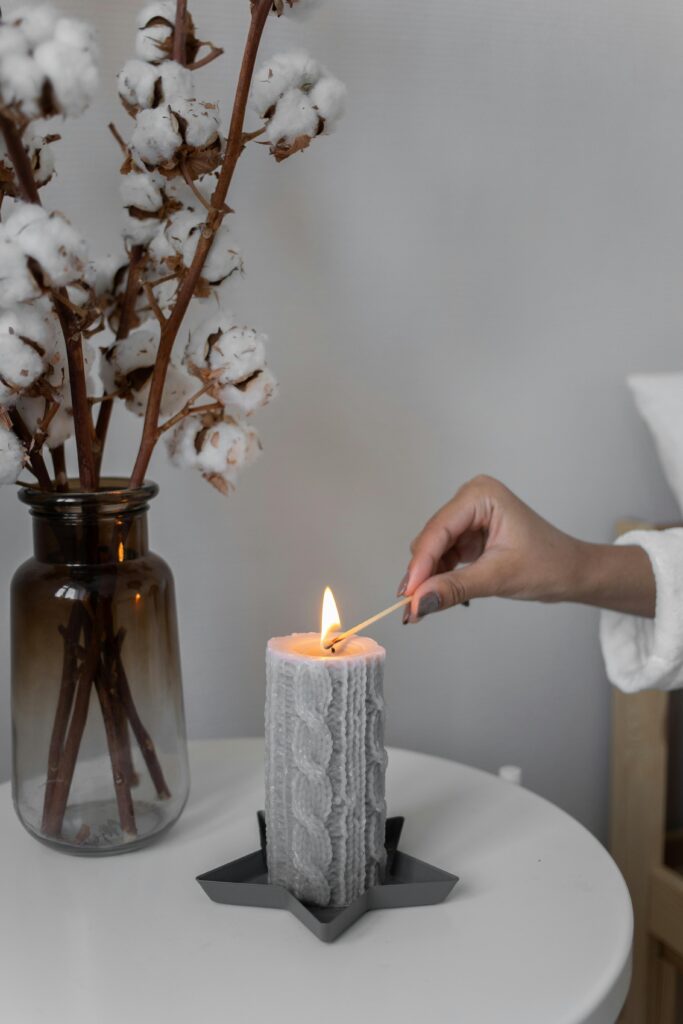 Ambiente com vela grande lindamente decorada sendo acessa. Ao lado um vaso de vidro fumê com galhos de algodão.