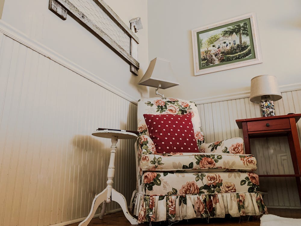 Cantinho da casa com estilo de decoração vintage: poltrona antiga floral, almofada poá, abajur com cúpula delicada e quadro com bela paisagem.