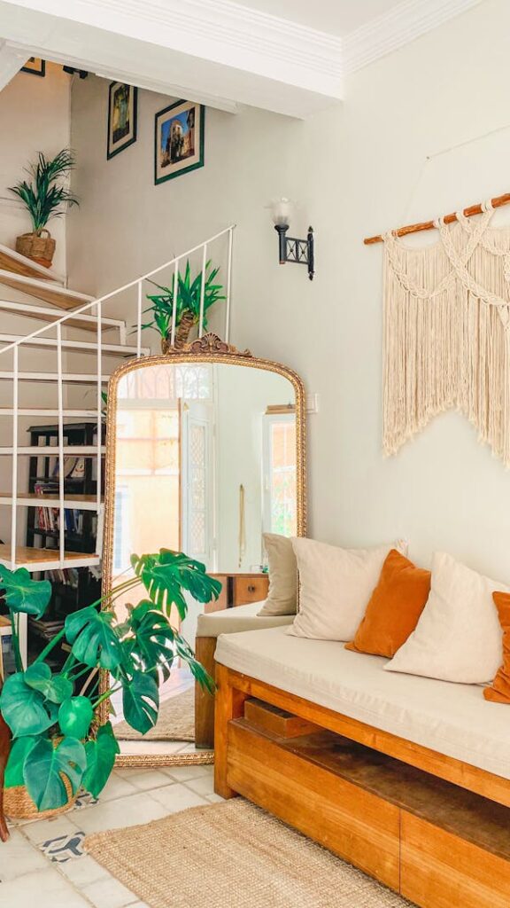 Sala com estilo boho na decoração: macramê na parede, plantas em cachepot, grande espelho, almofadas claras e em tom alaranjado.