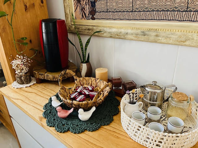 Foto dos Estilos de Cantinho do Café no estilo boho, com aparador em madeira clara, bandeja de palha, sousplat de crochê verde, utensílios artesanais, plantinhas e objetos afetivos.
