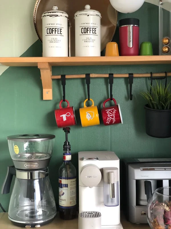 Cantinho do café em casa com parede verde, canecas coloridas penduradas e diferentes cafeteiras organizadas em bancada criativa.