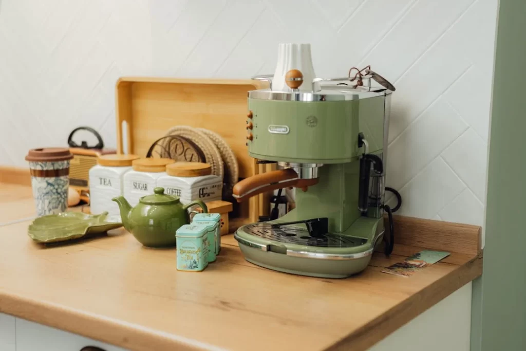 Cantinho do café em casa com cafeteira retrô verde, potes de cerâmica e bule artesanal sobre bancada de madeira clara.