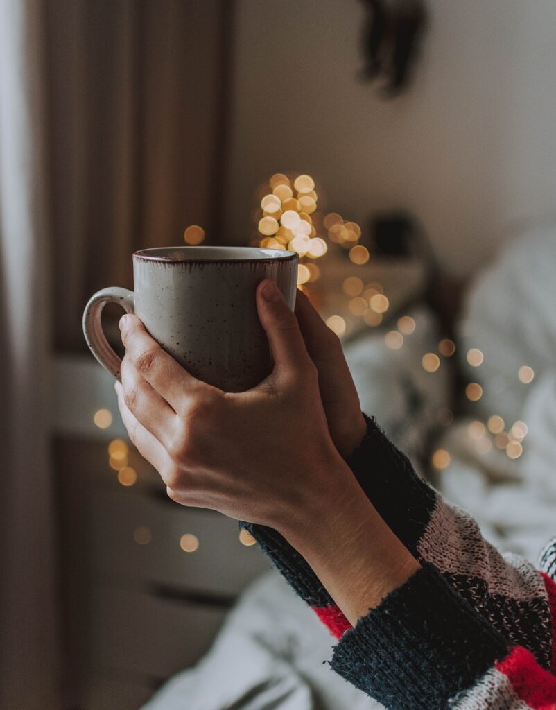 Cantinho do café em casa em momento de pausa, com mão segurando uma caneca em ambiente aconchegante com luzes suaves ao fundo.