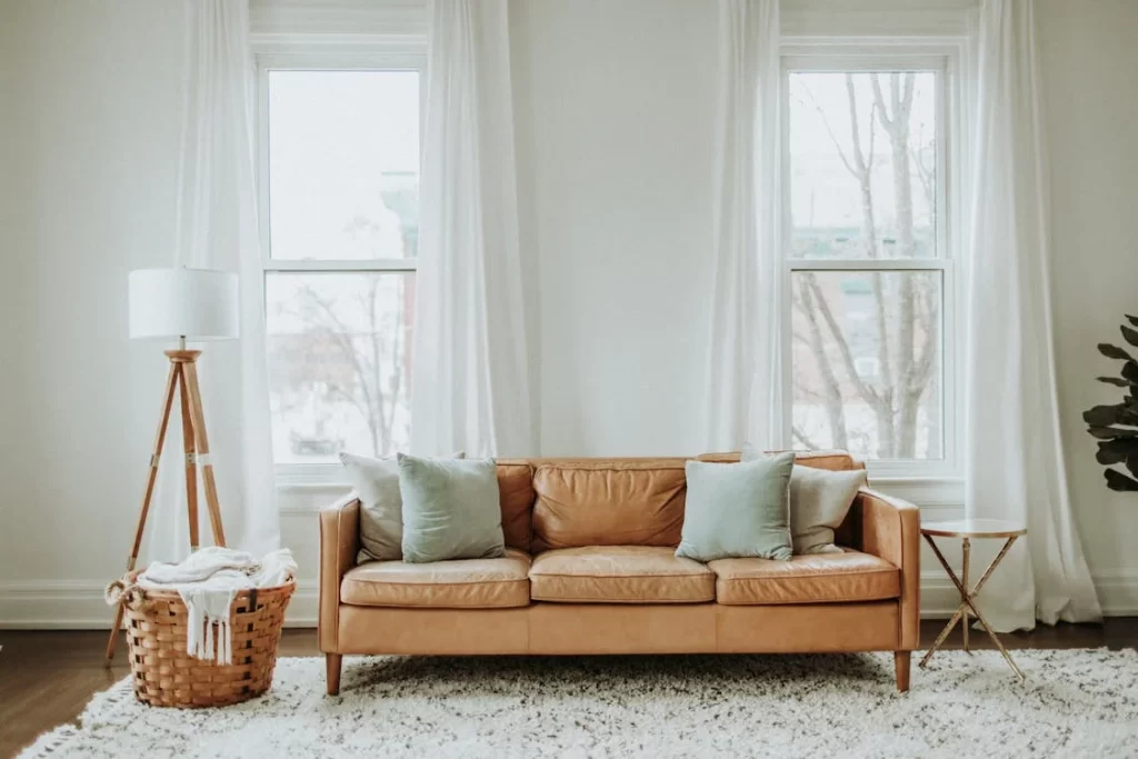 Sala de visitas com estilo boho na decoração: sofá de couro bege, cesta de palha com manta, planta, tapete claro com franjas, almofadas cinza claro.