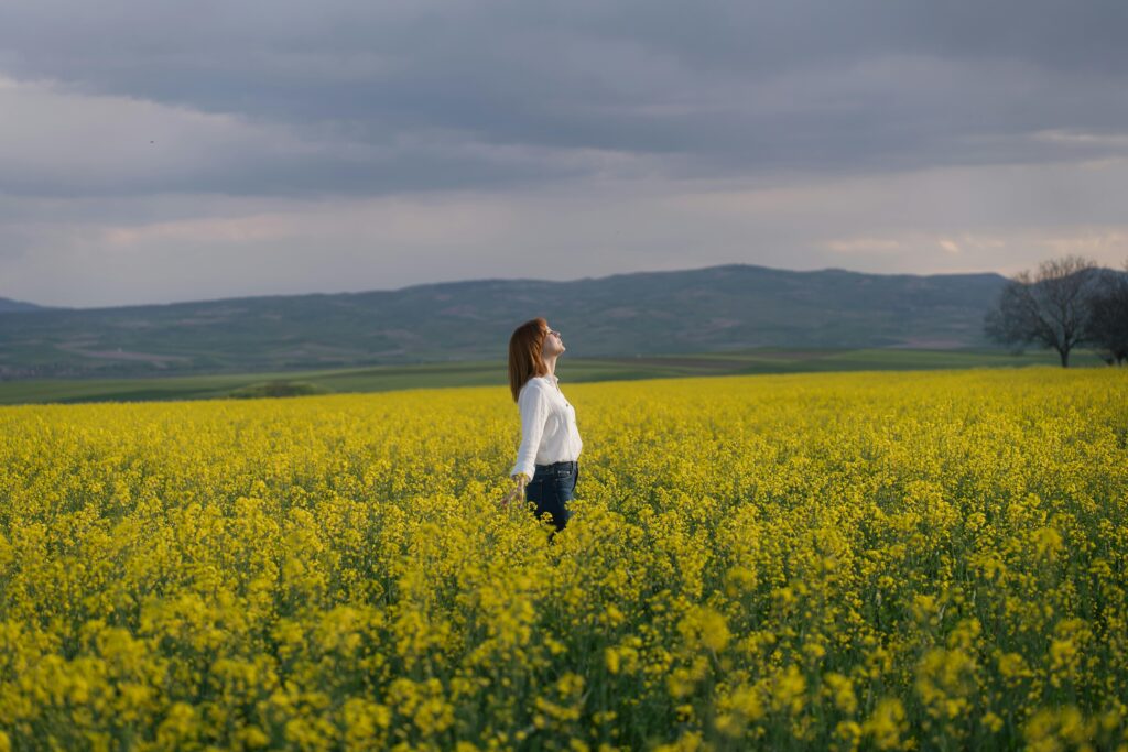 Mulher de braços abertos contemplando um campo florido sob o céu nublado.