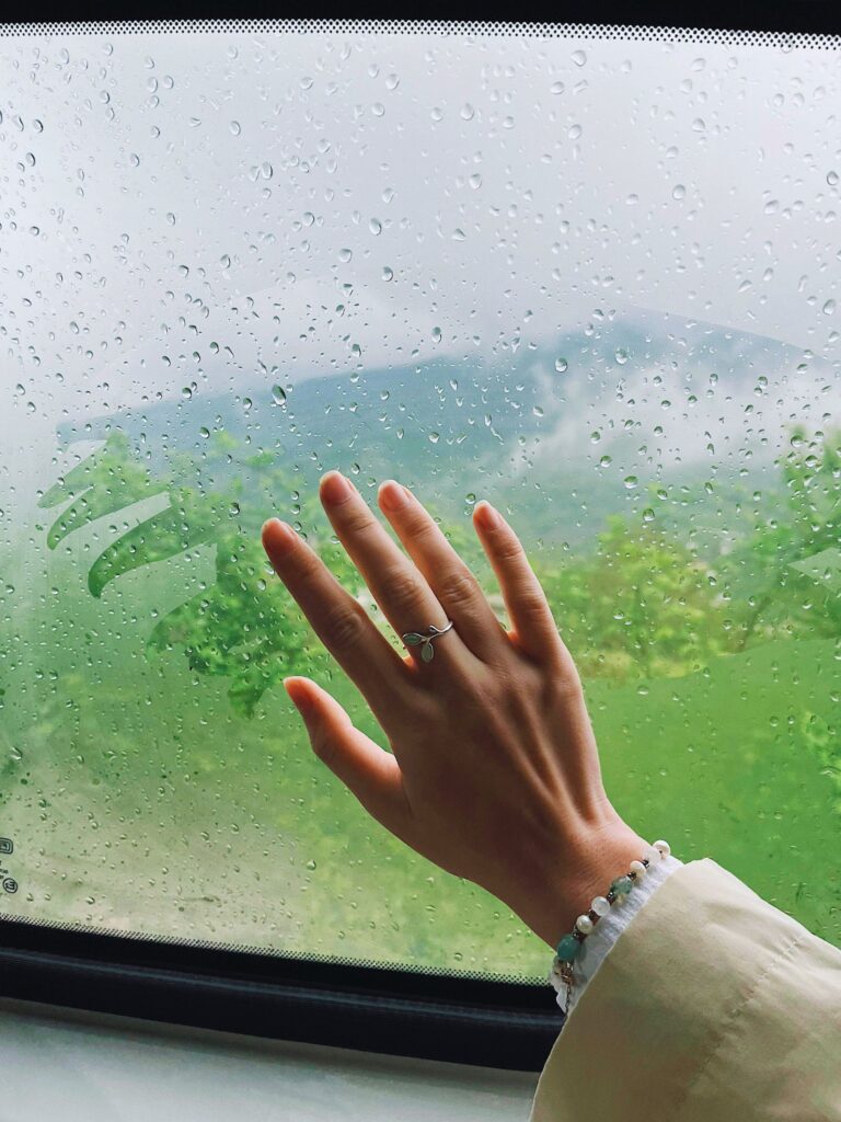 Mão tocando janela com chuva, refletindo sobre o tempo que floresce e a confiança em Deus.