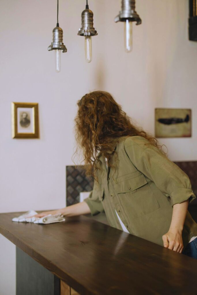 Mulher limpando uma mesa de madeira com pano, em ambiente simples e acolhedor.