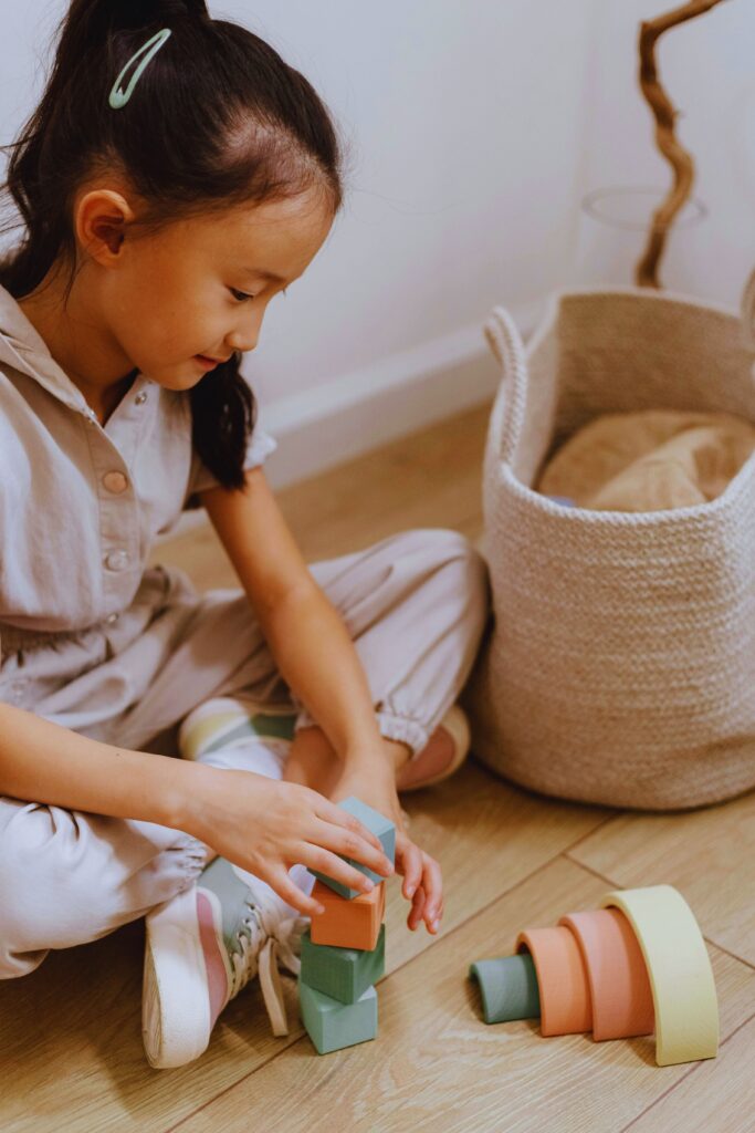 Organização da sala com crianças pequenas, menina brincando com blocos coloridos ao lado de cesto de tecido.