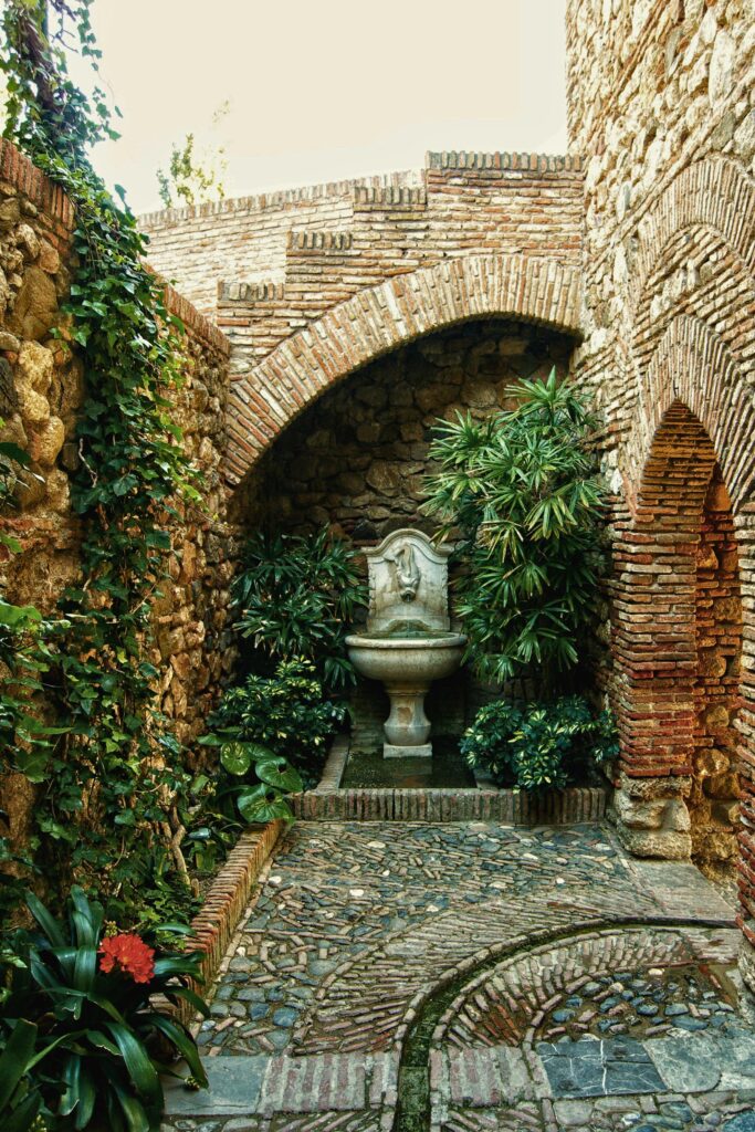 Fonte de água decorativa em pátio de pedra com arcos e vegetação ao redor