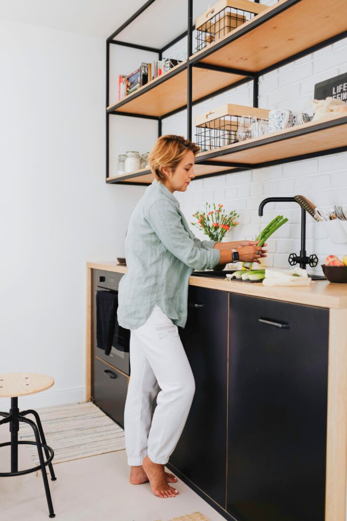 Mulher cortando legumes em cozinha moderna com armários pretos e prateleiras industriais.